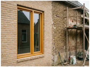 Nieuw houten kozijn in Nederlandse bakstenen gevel naast verweerde muur in renovatie, warme eikentinten en zandkleurig metselwerk.